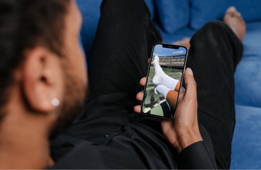 A man in a black shirt and pants reclines on a blue couch while holding a smartphone that displays a photo of white knit socks.